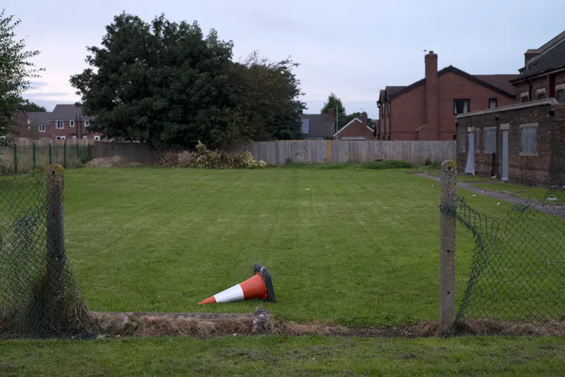 An overturned construction cone next to a missing segment of chain linke fence in Warrington, England.
