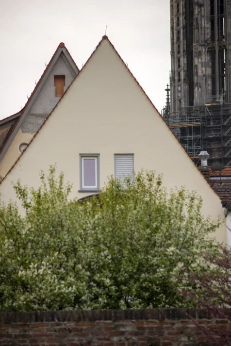 A pair of buildings, bush, brick wall and Gothic cathedral in the background in Ulm, Germany, 2023.