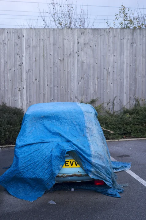 A partially-covered Mini Cooper is parked next to a wooden fence in Warrington, England, 2017.